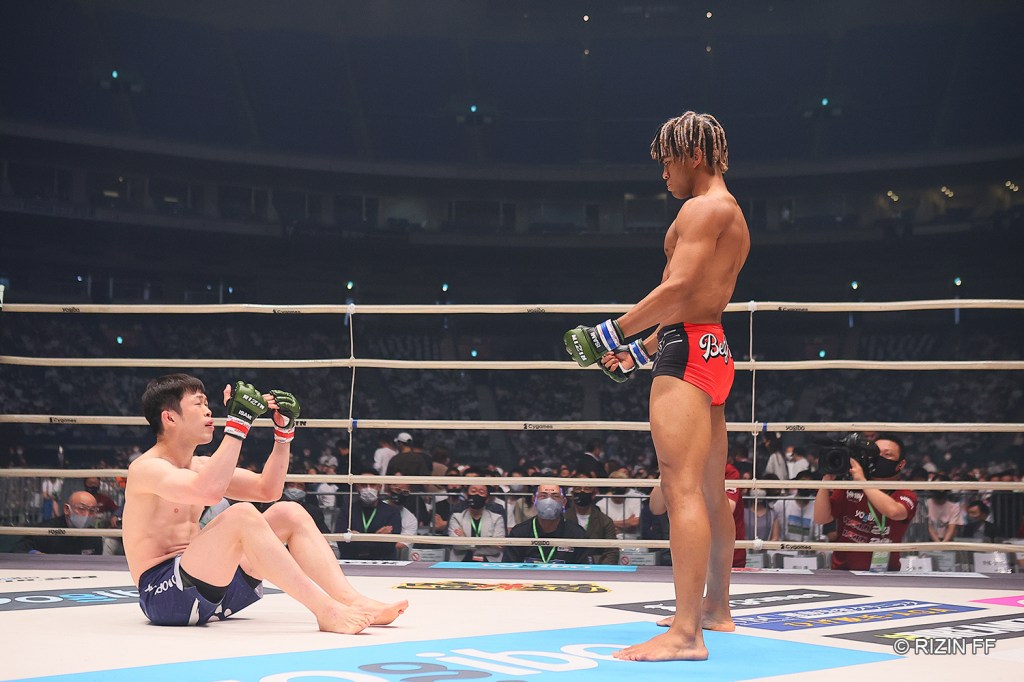 Satoshi Yamasu pleads while sitting in the RIZIN ring for BeyNoah to go to the ground for the fight. BeyNoah stands with his arms at his sides refusing to go down.