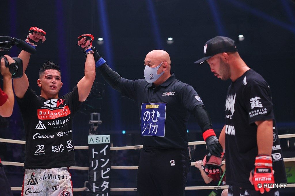 The referee raises the arm of Alan Yamaniha while Kazuma Kuramoto stands on his other side.