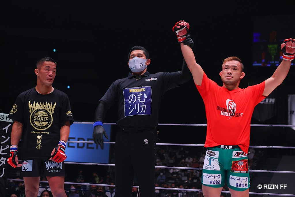 Hiromasa Ougikubo gets his arm raised by a referee during a decision announcement. On the other side of the referee, Takafumi Otsuka stands without his arm raised in victory.