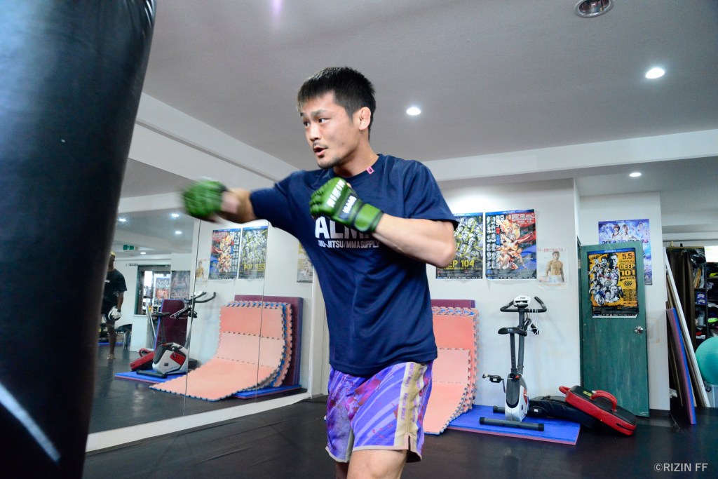 Juntaro Ushiku throws his right hand into a heavy bag inside a small MMA gym with a short roof.