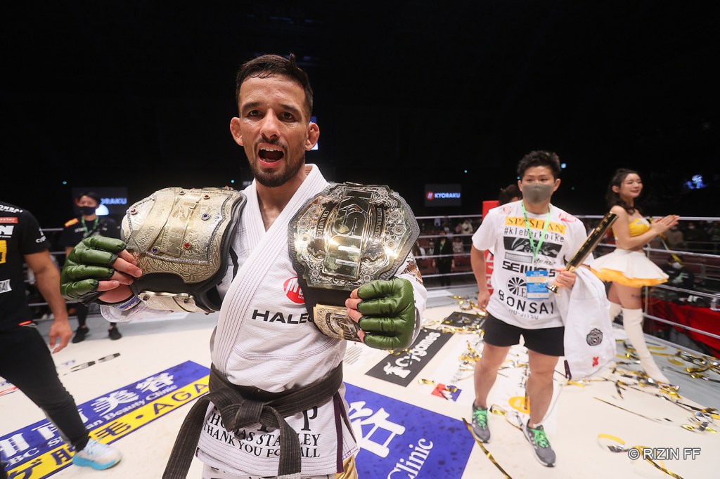 Kleber Koike holds a RIZIN and KSW belt on each shoulder while in the RIZIN ring. Cornerman and fighter Hiroaki Suzuki stands in the background.